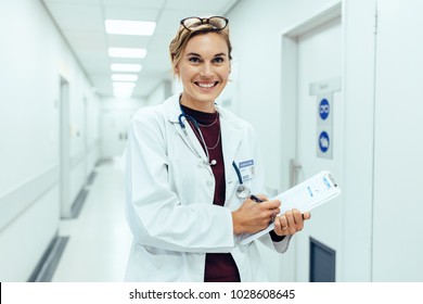 Portrait of happy young female doctor standing in hospital corridor and writing medical report on clipboard. Caucasian woman working in nursing home.