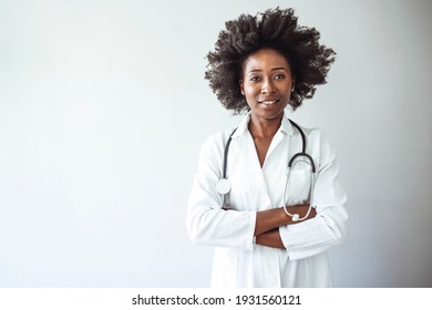 Portrait Of Happy Young Female Doctor Standing In Hospital Corridor. African Woman Working In Healthcare Center. Portrait Of Female Doctor Against Wall In Hospital