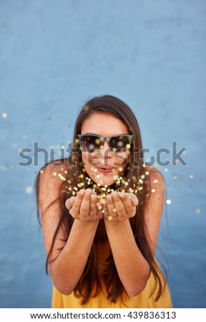 Portrait of happy young female model blowing confetti in the air over shining background