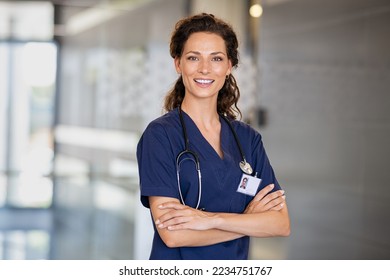 Portrait of happy young female nurse with folded arms standing in hospital hallway. Confident doctor woman in uniform and stethoscope looking at camera with copy space. Young healthcare worker working - Powered by Shutterstock