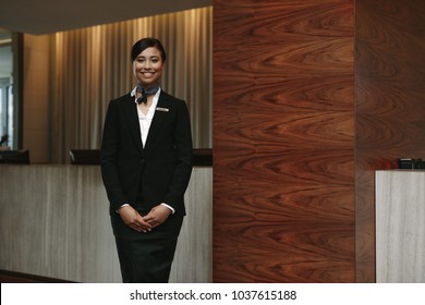 Portrait Of Happy Young Female Receptionist Standing At Hotel Front Desk To Welcome Guest. Female Concierge Welcoming Guest In Hotel.