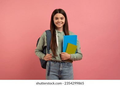 Portrait of happy young female student with backpack and books posing over pink studio background, beautiful teen girl holding workbooks and smiling at camera, enjoying modern education, copy space