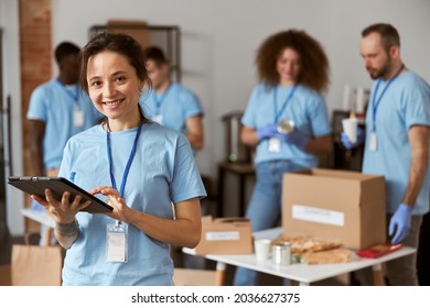 Portrait of happy young female volunteer in blue uniform using tablet pc and smiling at camera while standing indoors. Team sorting, packing items in the background - Powered by Shutterstock