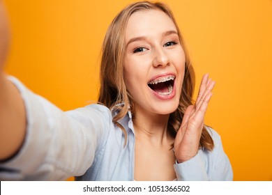 Portrait of a happy young girl with braces taking a selfie and laughing isolated over yellow background