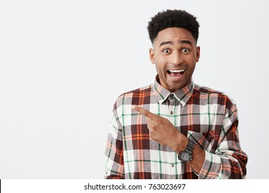 Portrait Of Happy Young Good-looking Tan-skinned Male Student With Afro Hairstyle In Casual Checkered Shirt Smiling, Pointing Aside With Finger, Looking In Camera With Excited Face Expression