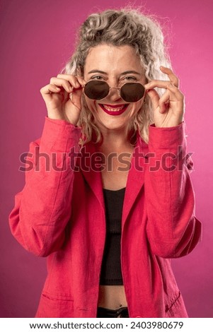 Portrait of a happy young hipster woman in a pink jacket isolated on a pink background leaning on a high chair with glasses