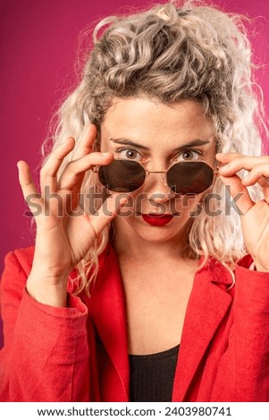 Portrait of a happy young hipster woman in a pink jacket isolated on a pink background leaning on a high chair with glasses