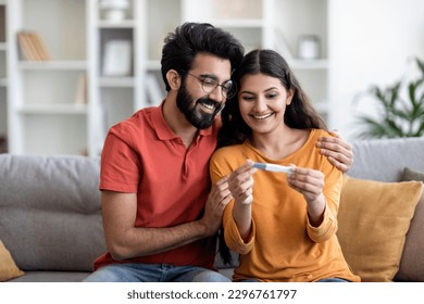 Portrait Of Happy Young Indian Couple Holding Positive Pregnancy Test While Sitting On Couch At Home Together, Eastern Husband Embracing His Joyful Wife, Celebrating Awaiting Baby, Copy Space