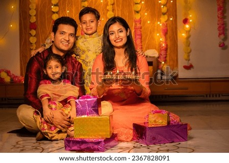 Portrait of happy young Indian family in traditional dress with lots of gifts around sitting on floor celebrating diwali festival at home.