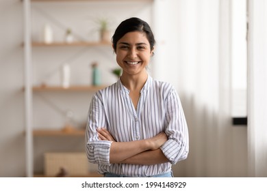 Portrait of happy young Indian female renter tenant stand arms crossed pose in living room at home. Smiling millennial successful confident mixed race ethnicity woman show leadership at workplace.