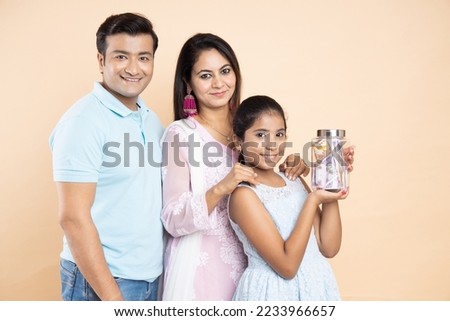 Portrait of happy young Indian parents with daughter holding glass jar full of rupee notes isolated on beige studio background. Saving money and investing concept.