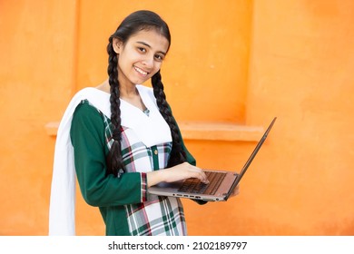 Portrait of Happy young indian school girl using laptop. Smiling braided hair female kid holding computer against orange background, She is looking at camera, skill india concept. - Shutterstock ID 2102189797