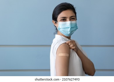 Portrait of happy young Indian woman in protective facemask rolling up sleeves, showing place with patch after covid vaccination prick, posing on grey wall, quarantine measures, healthcare concept.