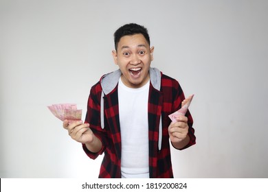 Portrait Of Happy Young Indonesian Man Holding Rupiah Money, Smiling Laughing Winning Gesture, Over White Background