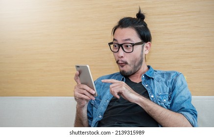 Portrait Of Happy Young Long Hair Nerd Asian Man Wearing Glasses Typing Sms On Smart Phone Lifestyle In Living Room. Technology Communication With Friends Family Work, Fun And Surprise Concept