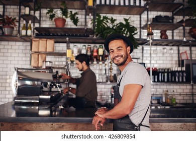 Portrait Of Happy Young Male Coffee Shop Owner Standing With Barista Working Behind The Counter Making Drinks.