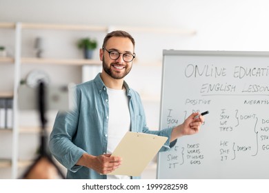 Portrait of happy young male teacher standing near blackboard with English grammar rules, conducting internet lesson. Positive tutor giving oreign language class on video call or web conference - Shutterstock ID 1999282598