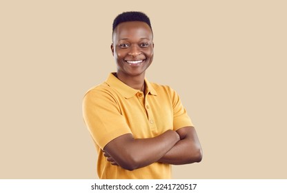 Retrato de un joven feliz con ropa informal. Alegre y guapo afeitado afroamericano con una camiseta de color amarillo arena, de pie con los brazos doblados sobre fondo beige, mirando la cámara y sonriendo