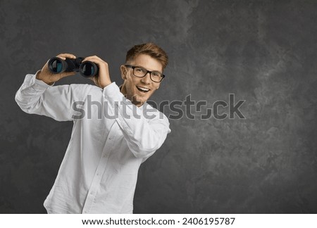 Portrait of happy young man in glasses holding optical zoom binoculars looking ahead standing against text copyspace studio background. College student searching for job or discovering something new