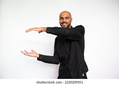 Portrait Of Happy Young Man Holding Invisible Object. Excited Latin American Male Model With Bald Head In Black Shirt Looking At Camera, Smiling While Showing Ads. Advertisement, Size Concept