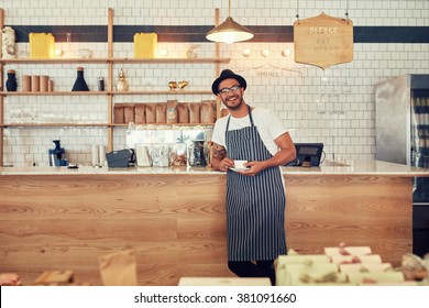 Portrait Of Happy Young Man Wearing An Apron And Hat Standing At A Cafe Counter Holding A Cup Of Coffee. Coffee Shop Owner Looking At A Camera And Smiling.