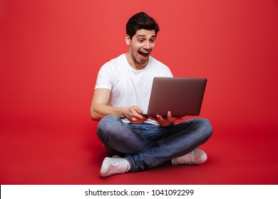 Portrait Of A Happy Young Man In White T-shirt Looking At Laptop Computer While Sitting On A Floor And Celebrating Isolated Over Red Background