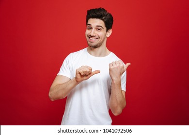 Portrait of a happy young man in white t-shirt pointing fingers away at copy space isolated over red background