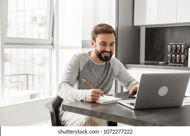 Portrait Of A Happy Young Man Working On Laptop Computer While Sitting At The Table At Home