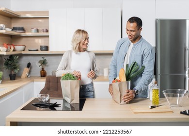 Portrait of happy young married couple coming home from shopping and unpacking paper bags with groceries in kitchen together. Household and relationship concept. - Powered by Shutterstock