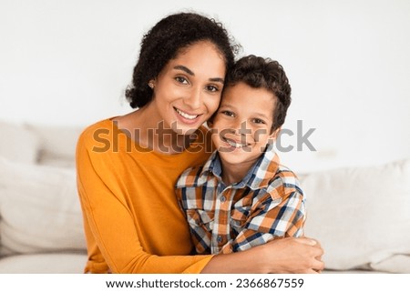 Portrait Of Happy Young Mom Embracing Her Kid Son Smiling To Camera At Home Interior. Boy Enjoying Mother's Hug Sitting On Couch Indoors. Family Bonding And Motherhood Concept