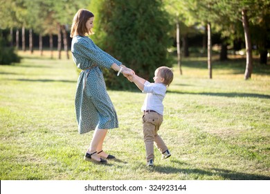 Portrait Of Happy Young Mom And Her Adorable Little Son Playing And Dancing Together In Park In Summertime, Smiling Mother And Kid Holding Hands, Spinning, Having Fun Together, Full Length