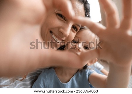 Portrait of happy young mother and small preschooler daughter look at camera posing making heart hand gesture, smiling millennial mom and little girl child show love and affection take selfie together
