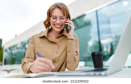 Portrait Of Happy Young Office Assistant Working Remotely Online Sitting With Laptop And Coffee Outside On Terrace Making Notes In Agenda In Same Time Speaking With Smile On Phone With Her Superior