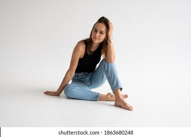 Portrait of happy young pensive woman posing in black underwear and blue jeans without a shoes, sitting on a white floor in white studio. Model tests of pretty girl in basic clothes on cyclorama.