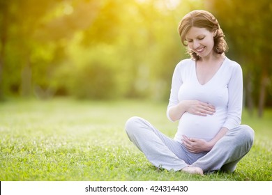 Portrait of happy young pregnant model sitting with crossed legs on grass lawn and looking at her belly with gentle smile. Future mom expecting baby caressing her tummy. Copy space