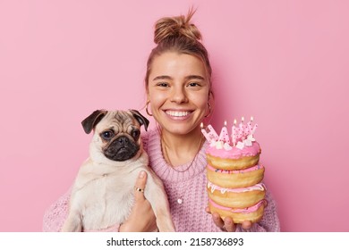 Portrait of happy young pretty woman poses with pug dog celebrates pets birthday holds pile of delicious doughnuts with burning candles isolated over pink background. Special occasion concept