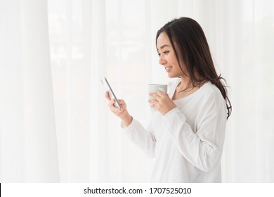 Portrait Of Happy Young Relax Asian Beautiful Woman Checking Social Media Online On Smartphone Or Mobile Phone While Drinking A Coffee And Stand In The Bedroom. Technology Communication Concept.