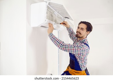 Portrait of happy young repairman fixing AC at work. Smiling handsome technician in workwear uses screwdriver to open up modern wall mounted air conditioner to check, repair it, or do disinfection - Shutterstock ID 2154465623