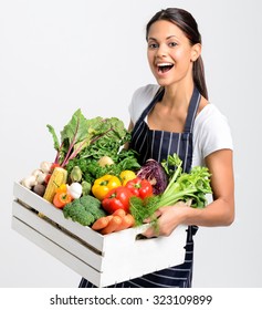 Portrait of happy young woman chef holding a crate full of fresh organic vegetables on grey background, promoting eating seasonally and sourcing from local producers