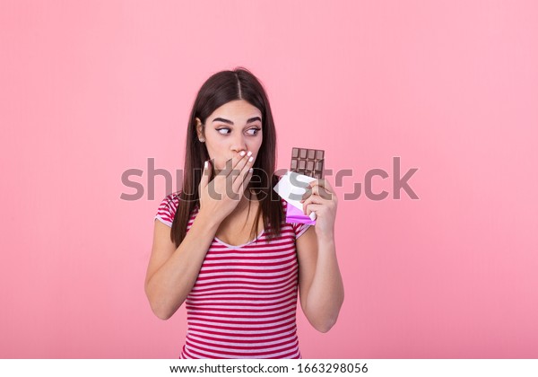 Portrait of a happy young woman with chocolate\
bar isolated over pink background covenring her mouth. Young woman\
with natural make up having fun and eating chocolate isolated on\
pink background