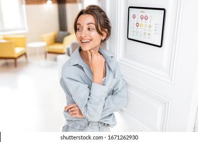 Portrait of a happy young woman controlling home with a digital touch screen panel installed on the wall in the living room