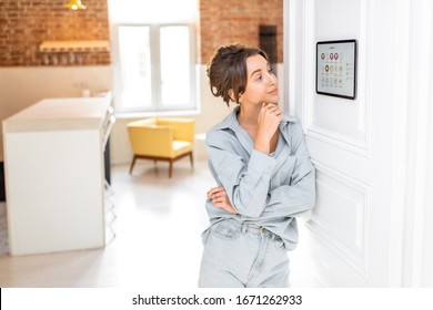 Portrait of a happy young woman controlling home with a digital touch screen panel installed on the wall in the living room