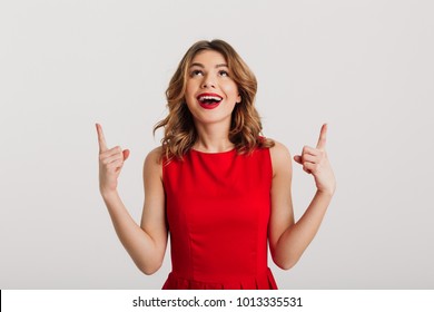 Portrait of a happy young woman dressed in red dress pointing fingers up at copy space isolated over white background