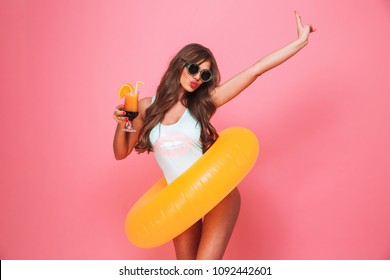 Portrait of a happy young woman dressed in swimsuit posing with inflatable ring and holding a cocktail isolated over pink background