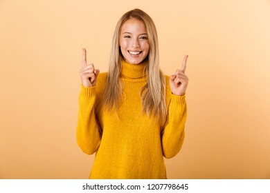 Portrait Of A Happy Young Woman Dressed In Sweater Standing Isolated Over Yellow Background, Pointing Up At Copy Space