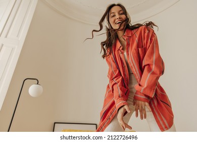 Portrait of happy young woman enjoys listening to music through headphones in bright room. Smiling beauty with wavy dark hair, in red shirt and white pants. Audio technology, gadgets and music concept - Powered by Shutterstock