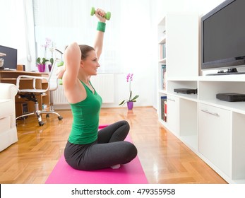 Portrait of a happy young woman exercising at home. She is sitting crossed-legged on the exercise mat in front of the TV and doing exercise for her arms with a pair of green hand weights. - Powered by Shutterstock