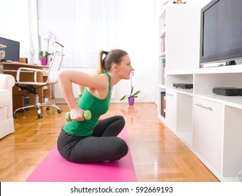 Portrait of a happy young woman exercising at home. She is sitting crossed-legged on the exercise mat in front of the TV and doing exercise for her arms with a pair of green hand weights. - Powered by Shutterstock