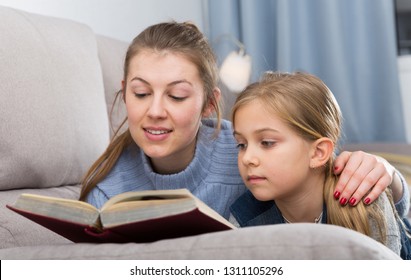 Portrait of happy young woman and her pre-teen daughter reading book at home