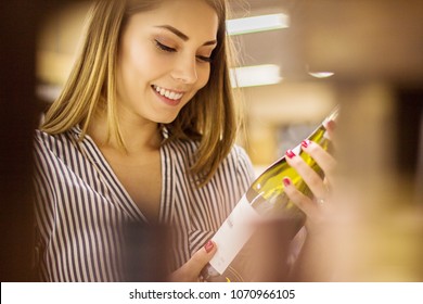 Portrait Of Happy Young Woman Holding White Wine Bottle In Hands And Smiling While Standing In Liquor Store. Beautiful Girl Looking At Blank Label With Copy Space While Shopping At Alcohol Supermarket